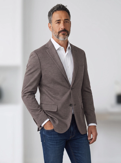 A man is standing indoors with one hand in his pocket and a neutral expression, wearing the 7 Downie St Alden Sport Coat Big & Tall by 7 Downie St in taupe over a white dress shirt and blue jeans.