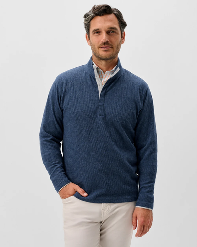 A man stands against a plain background wearing the Johnnie-O Ransel Hidden Snap Pullover Big & Tall in blue over a light shirt and beige pants, with one hand in his pocket.