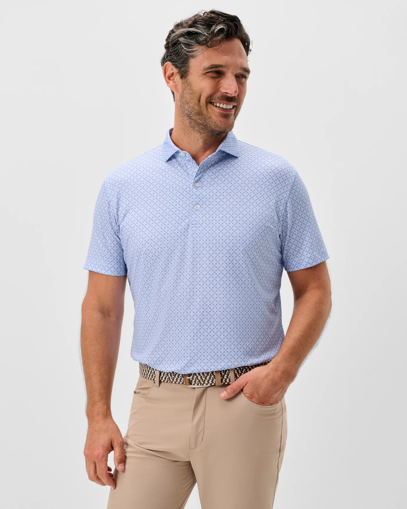 A man wears the Johnnie-O Torres Performance Big & Tall Polo, paired with beige pants and a patterned belt, standing against a plain light background.
