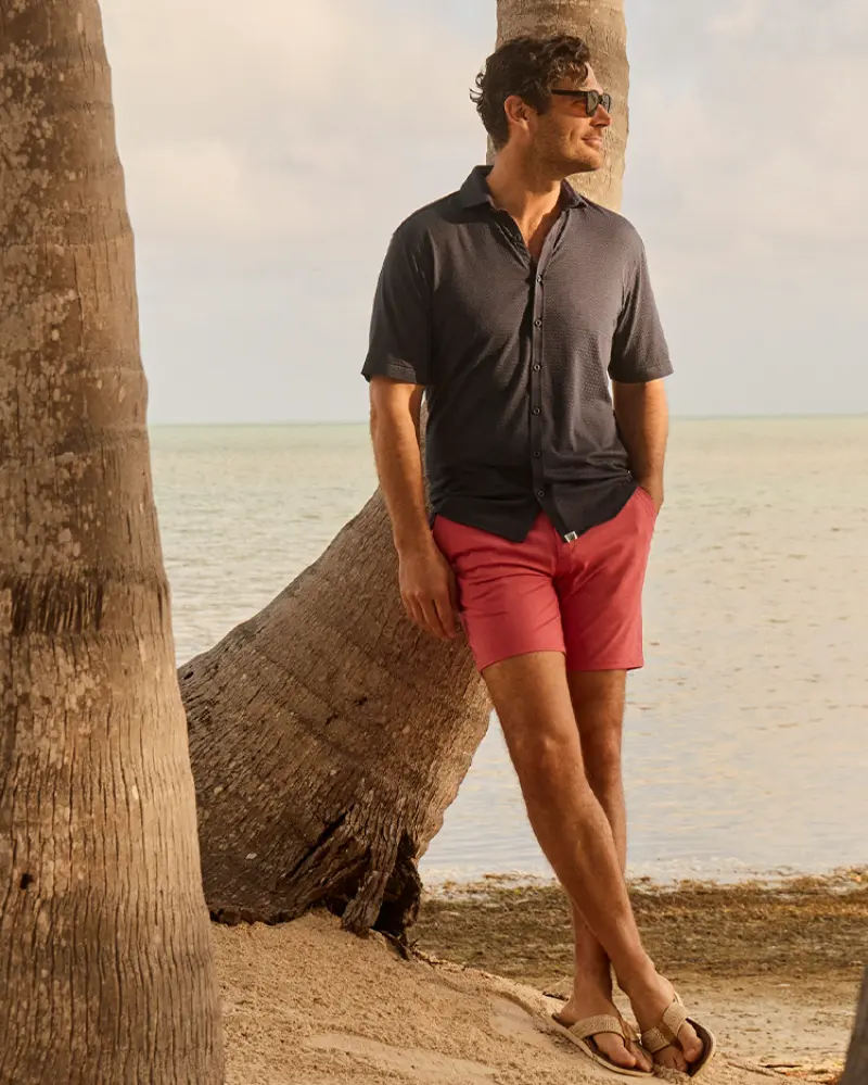 A man in a Johnnie-O Surfsucker Knit Short Sleeve Button Down Shirt leans against a palm tree on the beach, gazing at the water.