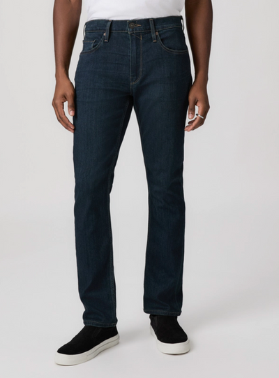 A person wearing Paige Federal Straight Slim Jeans by Paige in dark blue, paired with black slip-on shoes and a white top, is photographed from the shoulders down against a plain background.