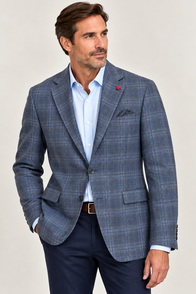 A man dressed in the TailoRed Giacomo Plaid Sport Coat 8110669—a blue plaid blazer by TailoRed—paired with a light blue dress shirt, navy pants, and a brown belt, stands with one hand in his pocket against a neutral background.