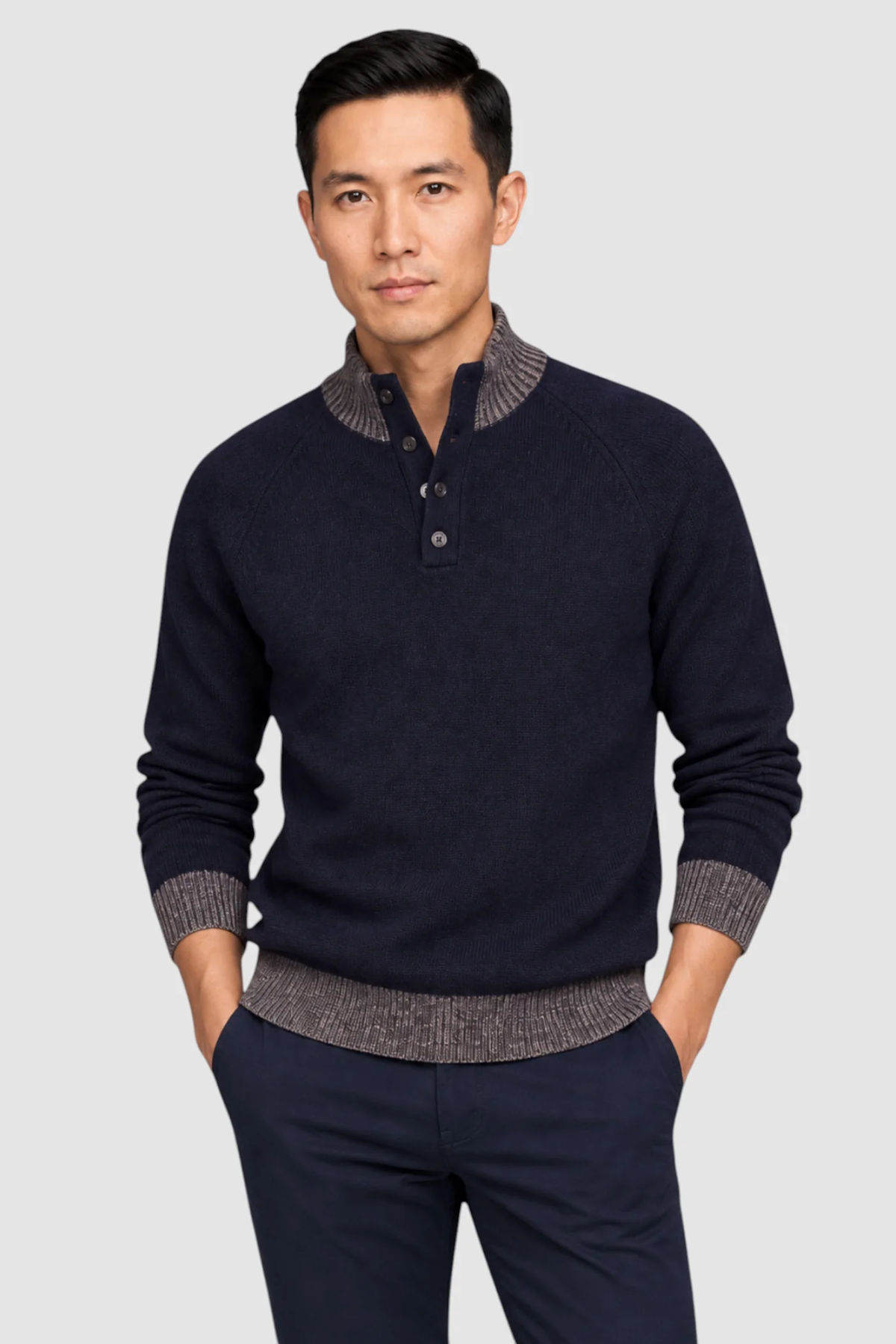 A man stands with his hands in his pockets against a plain light gray background, wearing the Garnet Tate Contrast-Trim Mock Neck Sweater 2622022 by Garnet, which features a refined navy body with contrasting brown collar, cuffs, and hem.