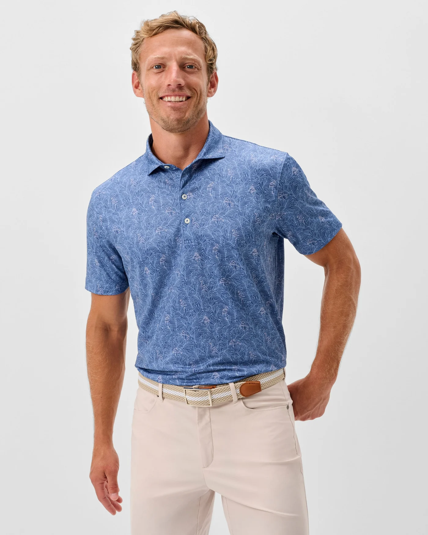 A man stands smiling in a Johnnie-O Osmond Big & Tall Polo by Johnnie-O, featuring moisture-wicking blue fabric, paired with light beige pants and a woven belt, set against a plain light background.
