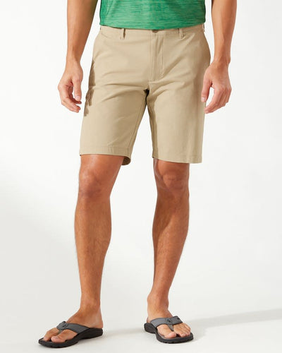A person wearing Tommy Bahama Chip Shot Shorts Big & Tall in tan, a green shirt, and black flip-flops stands against a plain white background.
