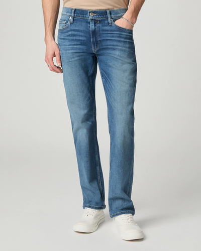 A person wearing Paige Federal Straight Slim Jeans by Paige in a medium wash with white sneakers stands with one hand in their pocket against a plain background.