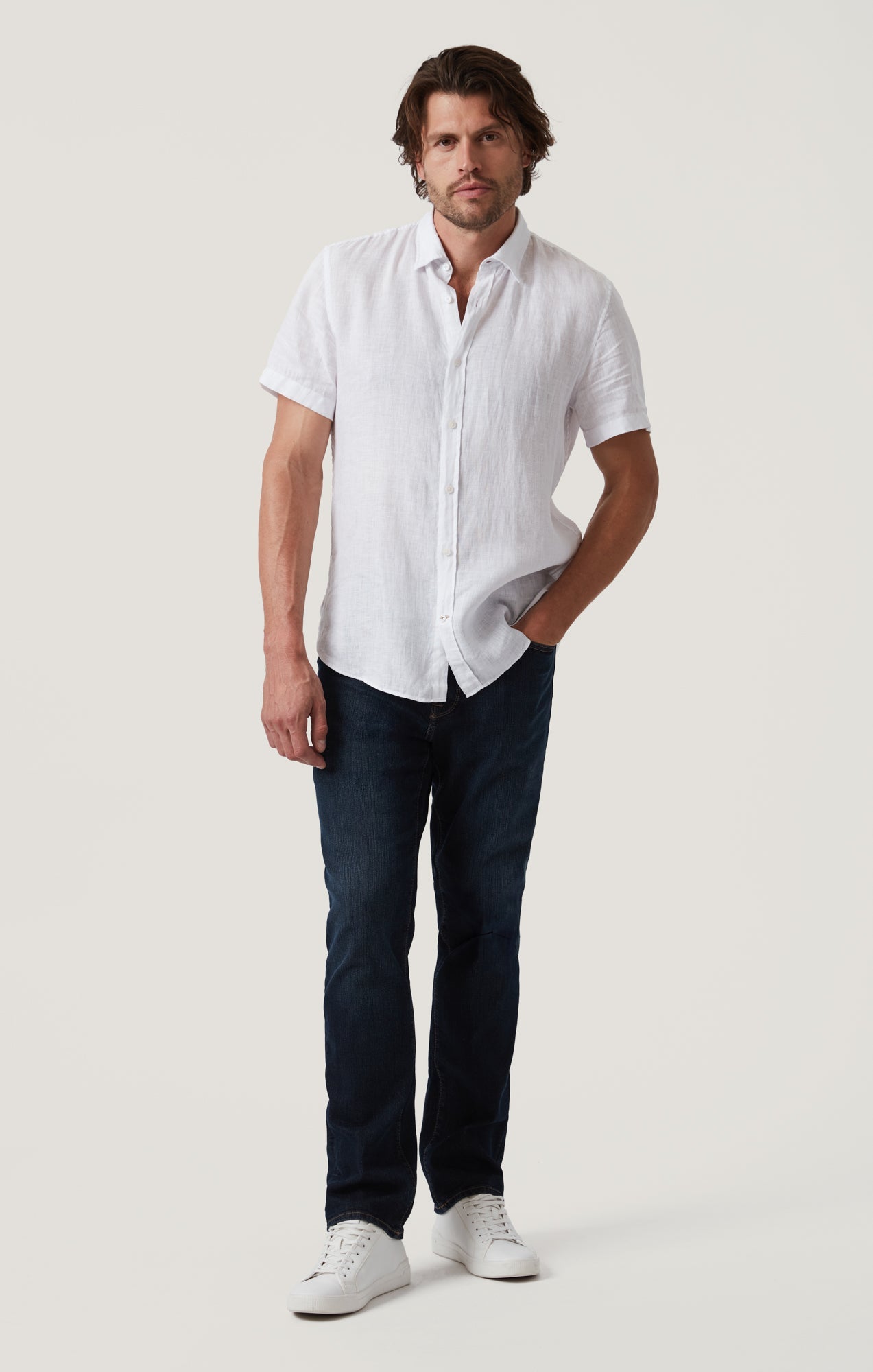 A man stands against a plain background wearing a white short-sleeve button-up shirt, 34 Heritage Dark Comfort higher-rise straight-leg dark blue denim pants, and white sneakers.