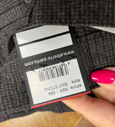 A hand holds an Alberto pants tag (alberto-pants.com) displaying Alberto Stone Mini Check Smart Wool Pants 1625, style 5007 Stone, with a barcode on wrinkle-resistant dark grey wool fabric.