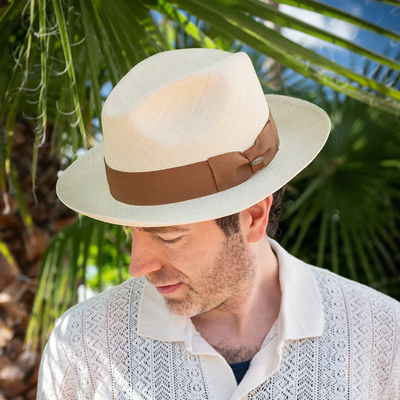 A man wearing the Scala Panama Safari - Roswell P219 hat by Dorfman Pacific, featuring a brown band, stands outdoors in a white patterned shirt near green palm leaves.