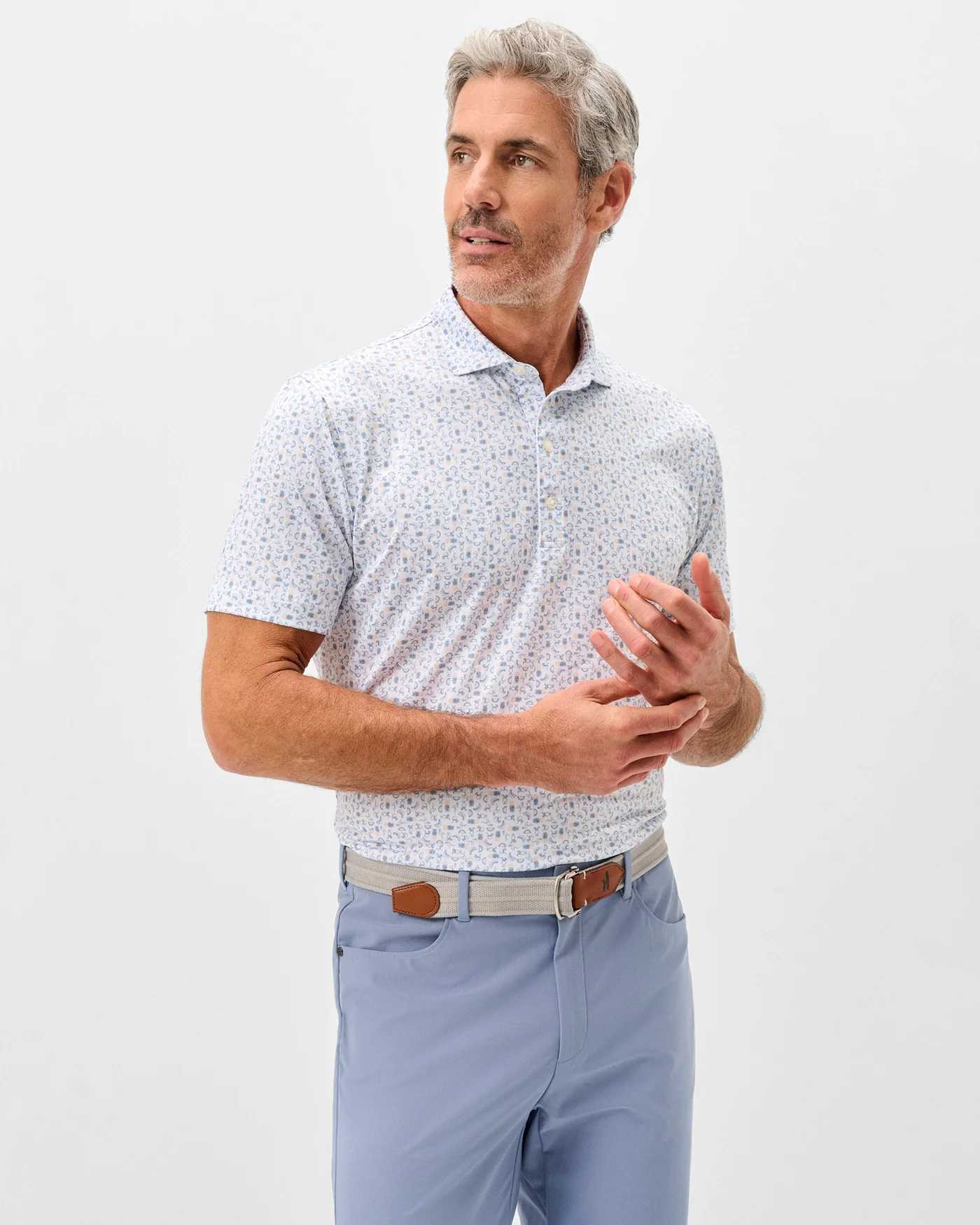 A man with gray hair and a beard wears the Johnnie-O Cabana Bar Performance Jersey Polo by Johnnie-O, styled with light blue trousers, standing against a plain white background.