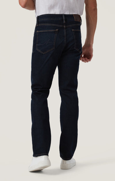 A person is shown from the back wearing 34 Heritage Dark Comfort jeans by 34 Heritage, featuring a straight leg fit in dark blue, paired with white sneakers; their right arm is bent while their left arm remains relaxed.
