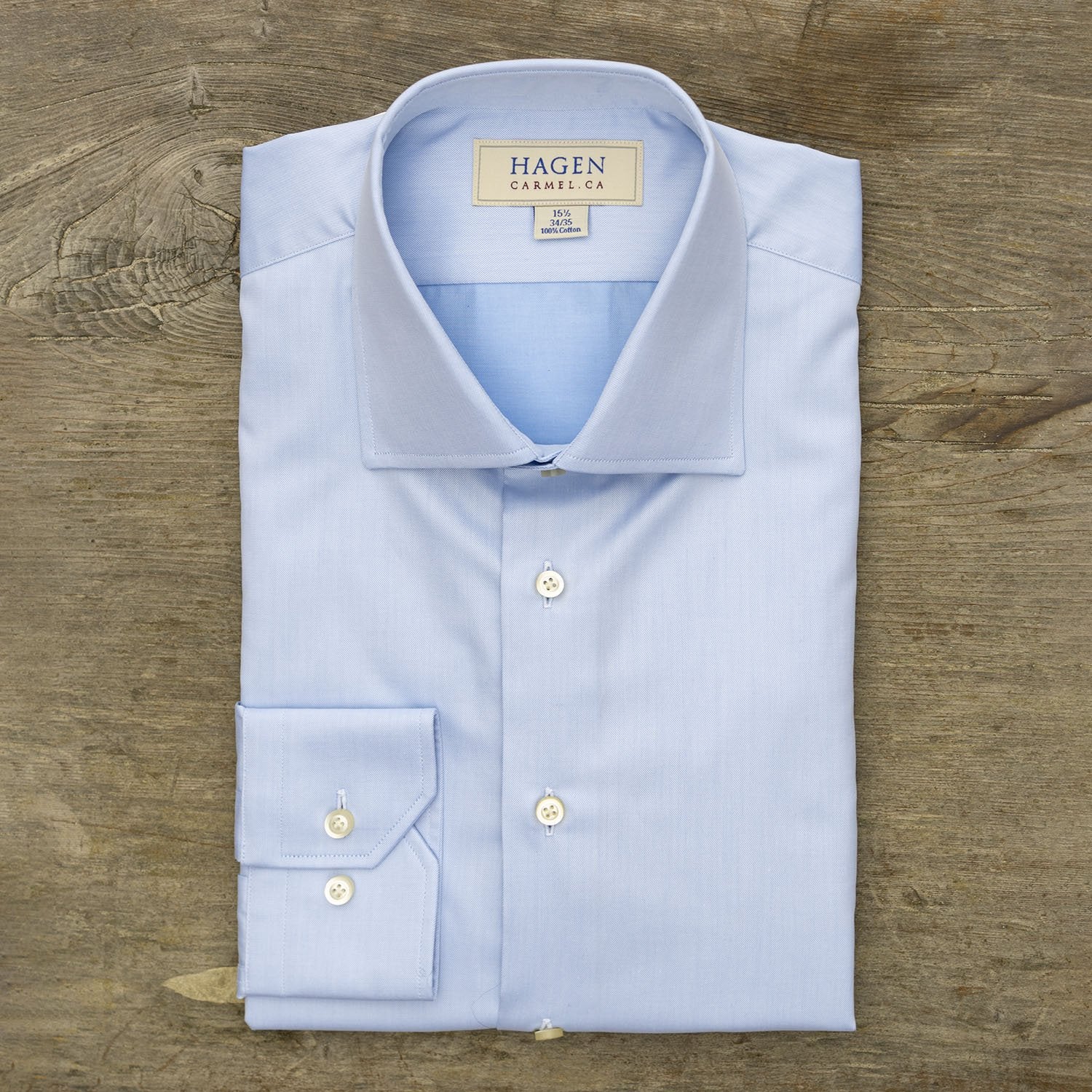 A neatly folded Hagen Solid Twill Shirt MB1864400, crafted from 100% cotton superfine twill with white buttons, is displayed on a wooden surface. The label reads Hagen, Carmel, CA.