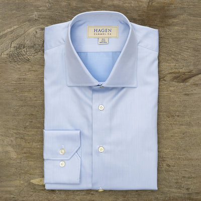 A neatly folded Hagen Solid Twill Shirt MB1864400, crafted from 100% cotton superfine twill with white buttons, is displayed on a wooden surface. The label reads Hagen, Carmel, CA.