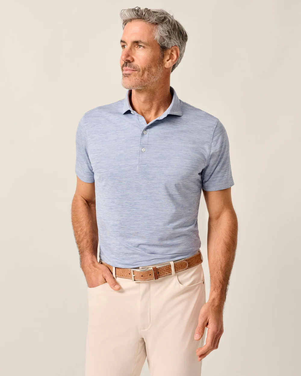 A man wearing a Johnnie-O Performance Polo-Huronn in light blue, beige pants, and a brown belt stands against a plain light background, looking to the side.