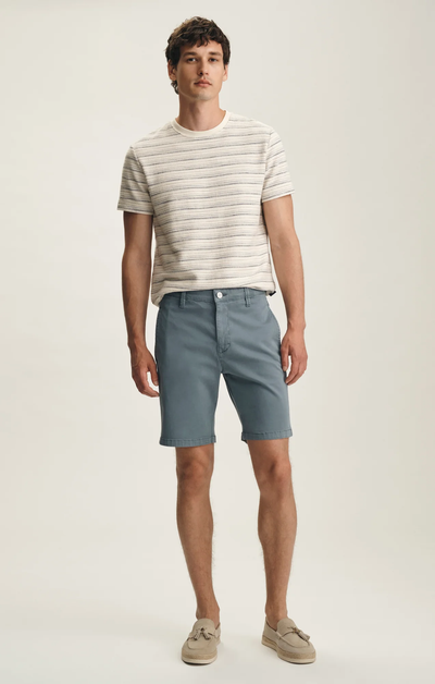 A man stands against a plain background wearing a short-sleeve striped shirt, 34 Heritage Nevada Twill Shorts in light blue made from breathable stretch fabric, and beige loafers.