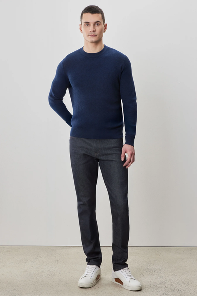 A man stands against a plain light wall, wearing the Robert Barakett Watertown Sweater by Robert Barakett, paired with dark jeans and white sneakers.