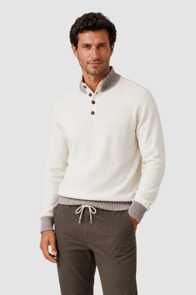 A man wears the Garnet Tate Contrast-Trim Mock Neck Sweater 2622022 by Garnet—a white long-sleeve knit style featuring a button collar and grey contrast cuffs—paired with dark drawstring pants, standing against a plain light background.
