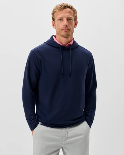A man stands against a plain background, wearing the Johnnie-O Willard Performance Hoodie Big & Tall in navy blue, styled over a pink collared shirt and light gray pants, with his hands in his pockets.
