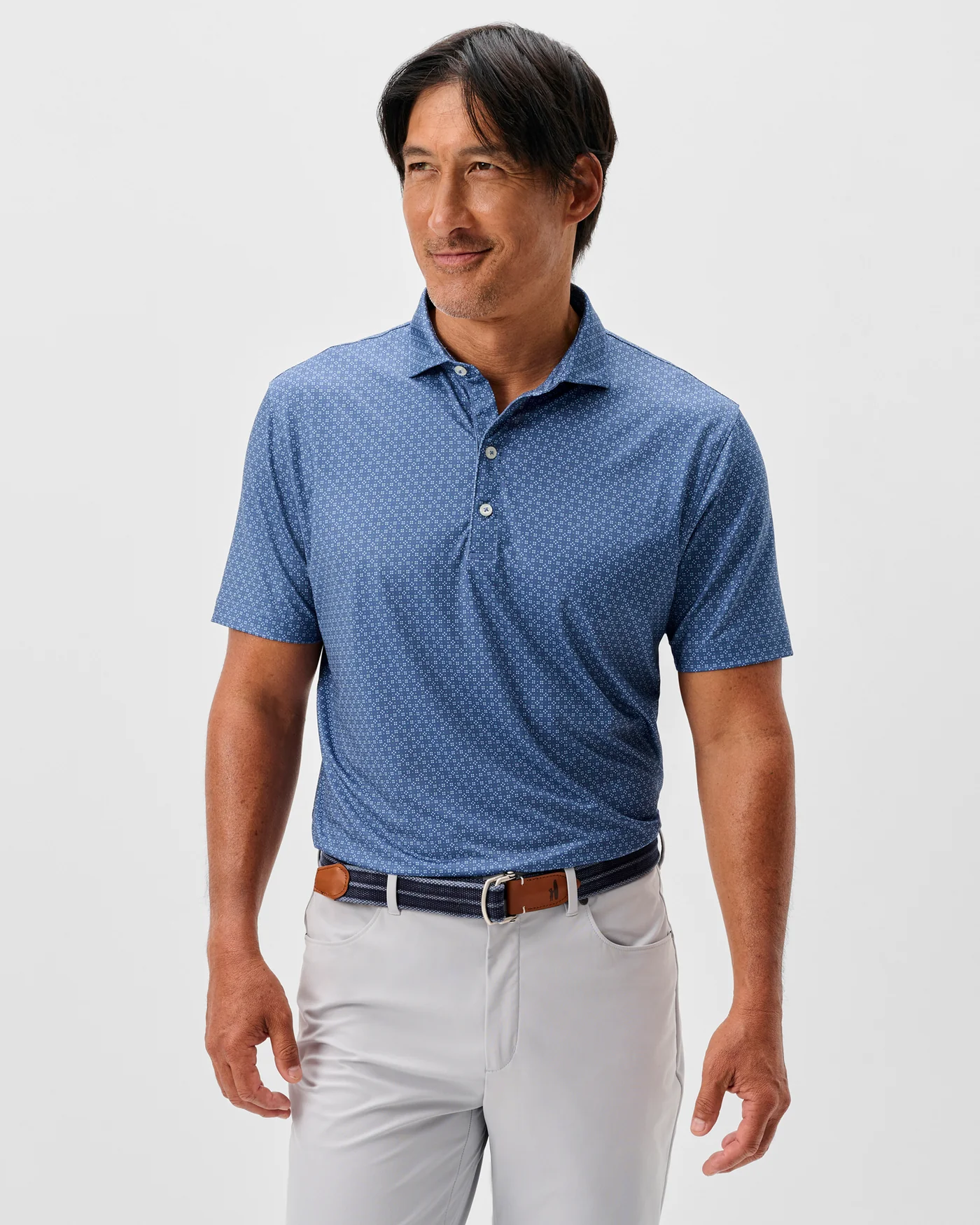 A man wears the Johnnie-O Torres Performance Big & Tall Polo tucked into light gray pants with a brown belt, standing against a plain light background.