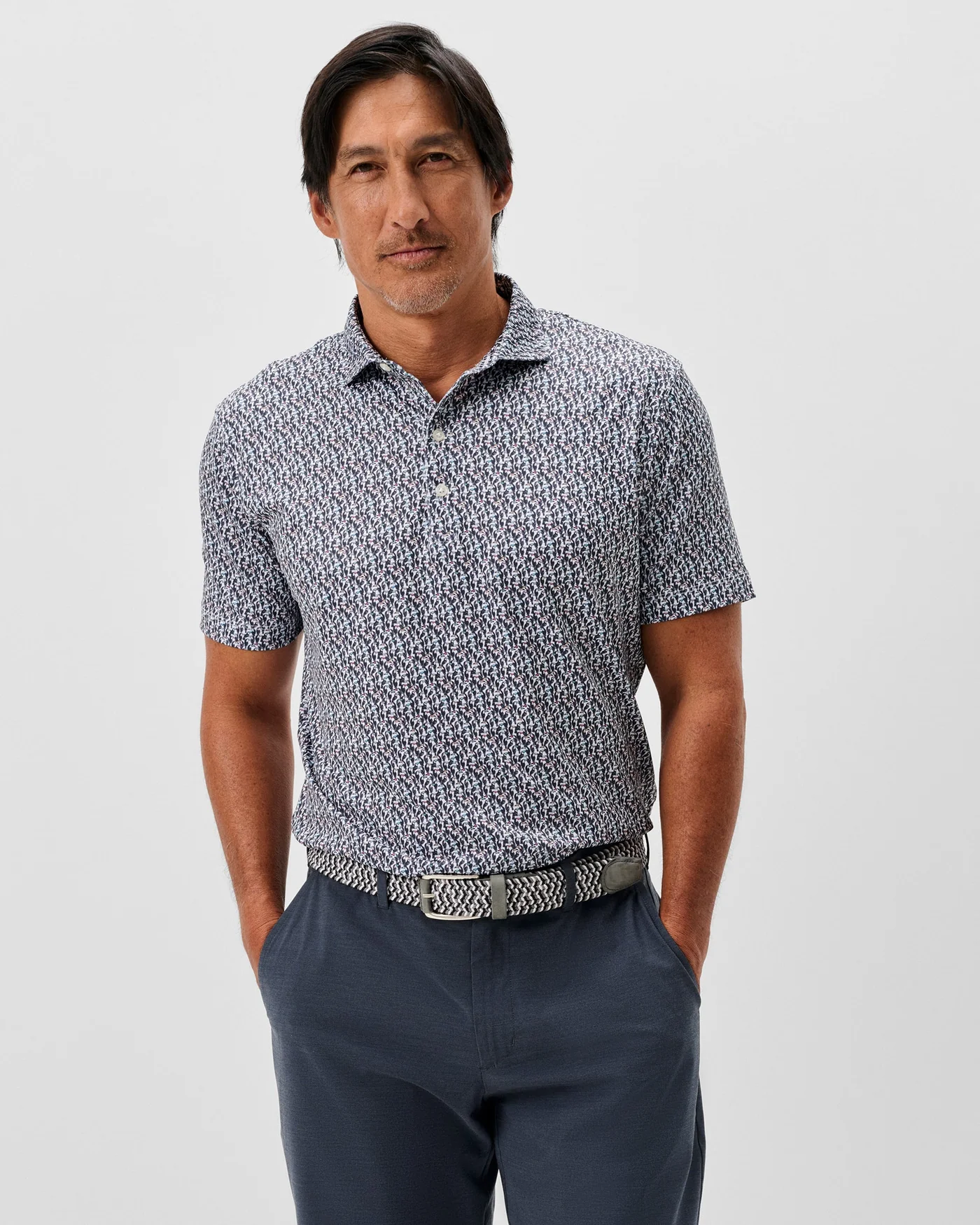 A man stands against a plain background with his hands in his pockets, wearing the Johnnie-O Toco Performance Jersey Polo, featuring short patterned sleeves, dark pants, and a patterned belt.