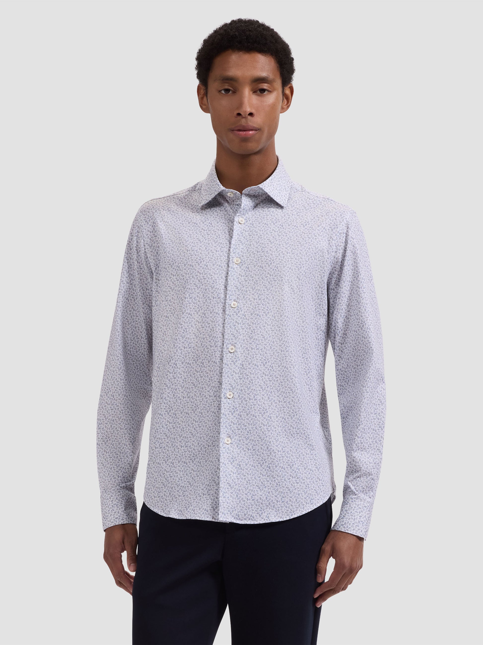 A man wears the Bugatchi OP James OoohCotton® Leaf Print Shirt JF9212K60, a light-colored, long-sleeve shirt with a subtle leaf print by Bugatchi, paired with dark pants against a plain light gray background.