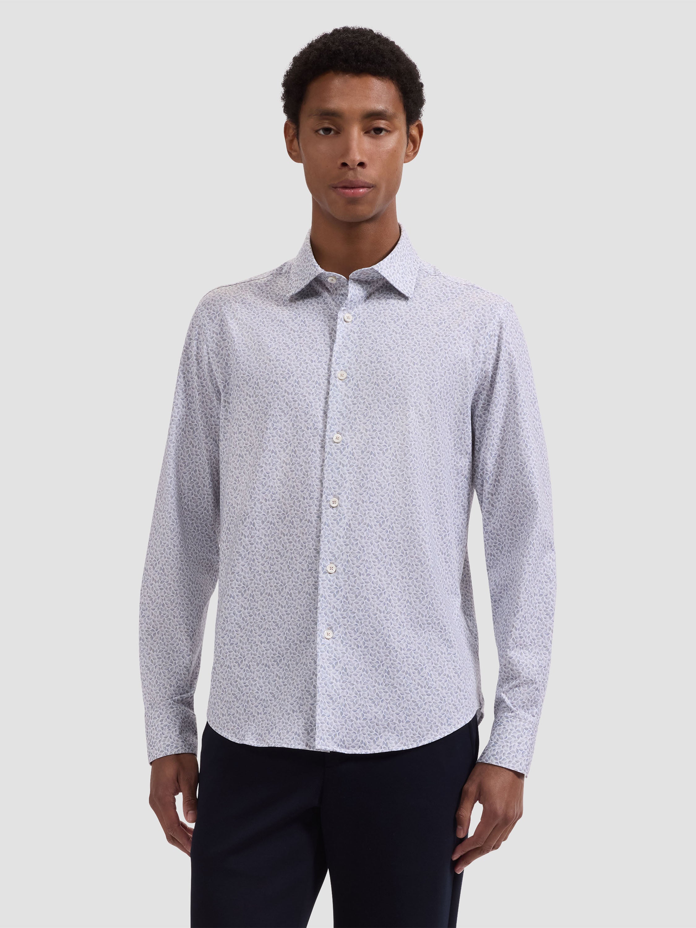 A man wears the Bugatchi OP James OoohCotton® Leaf Print Shirt JF9212K60, a light-colored, long-sleeve shirt with a subtle leaf print by Bugatchi, paired with dark pants against a plain light gray background.