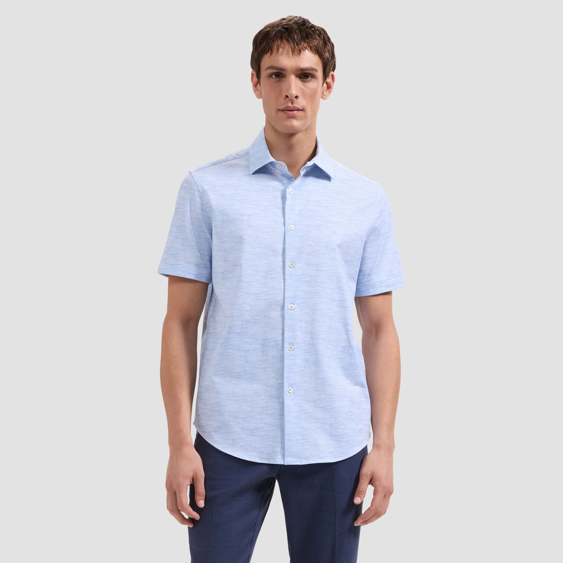 A man wears the Bugatchi Miles OoohCotton® Melange Shirt JF9505F40, a light blue 8-way stretch shirt by Bugatchi, with dark pants, standing against a plain light gray background.