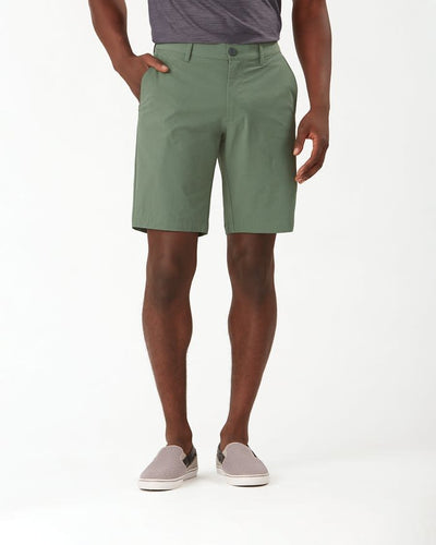A person wearing a gray t-shirt, Tommy Bahama Chip Shot Shorts Big & Tall in green with UPF 30 protection, and gray slip-on shoes stands with one hand in their pocket against a plain white background.