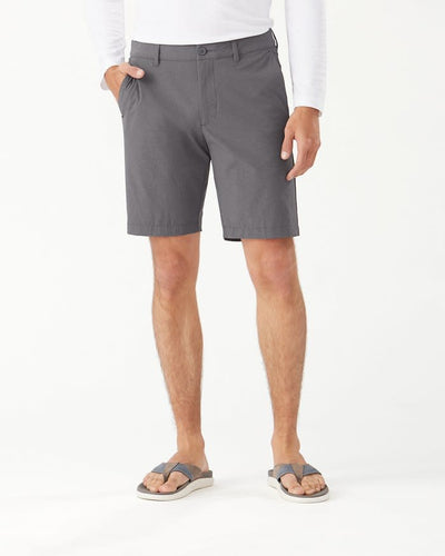 A person wears Tommy Bahama Chip Shot Shorts Big & Tall in gray, paired with a white long-sleeve shirt and gray sandals. The moisture-wicking shorts from Tommy Bahama offer comfort and UPF 30 sun protection against a plain white background.