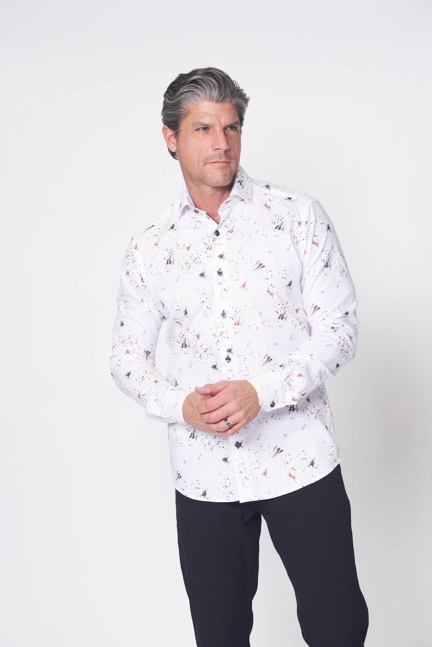 A man wearing the 7 Downie St. Big & Tall Sport Shirt White 27 LS by 7 Downie St, featuring a long-sleeve design and black and brown splatter pattern, paired with black pants, stands against a plain white background.