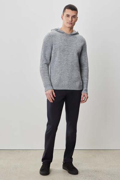 A man stands against a plain white wall, wearing the Robert Barakett Wildman Sweater by Robert Barakett, along with dark pants and black shoes.