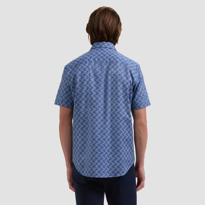 Person with brown hair shown from the back, wearing the Bugatchi Miles OoohCotton® Leaf Print Shirt JF9138F40—a short-sleeve, blue patterned performance shirt—paired with dark pants against a plain background.
