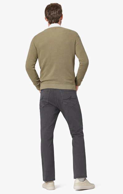A man stands facing away, wearing the 34 Heritage Charisma Diagonal Big&Tall dark gray jeans and a light olive sweater with white sneakers against a white background.