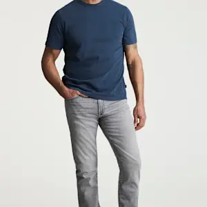 A man stands against a plain background, wearing a blue T-shirt and light gray 34 Heritage Courage Jeans Urban by 34 Heritage, with one hand in his pocket.
