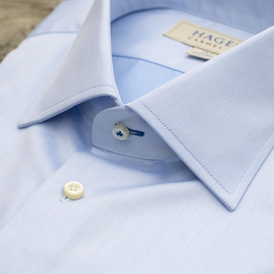 Close-up of the Hagen Solid Twill Shirt MB1864400 by Hagen—a neatly folded blue dress shirt made from superfine twill 100% cotton, featuring a pointed collar and white buttons, displayed on a wooden surface.