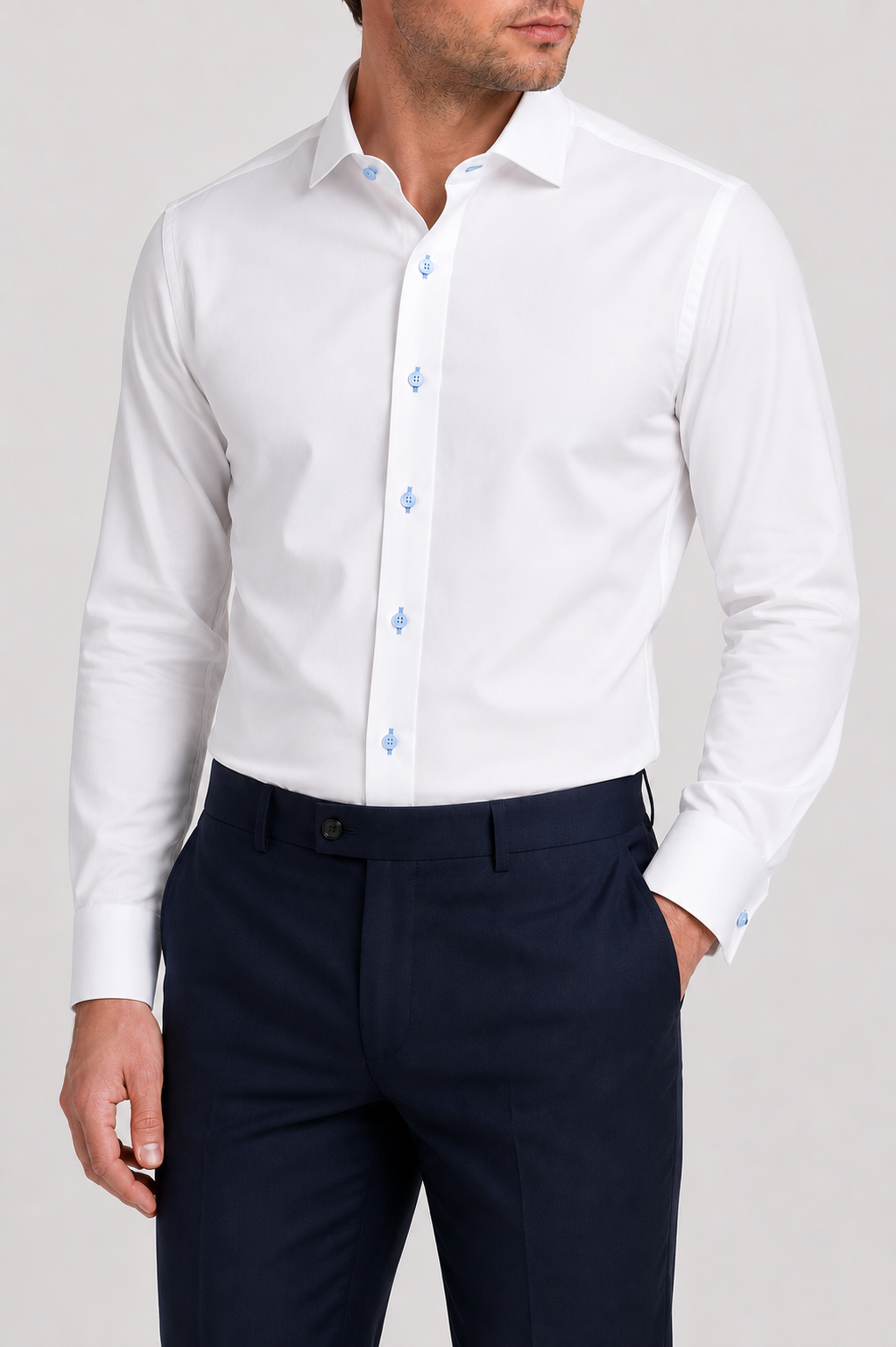 A man wearing the Wilkes & Riley White/Sky Spread Collar B1715 dress shirt, featuring a crisp white twill fabric with light blue buttons, pairs it with dark navy trousers. The refined spread collar from Wilkes & Riley adds sophistication to his polished look as he stands with one hand in his pocket against a plain background.