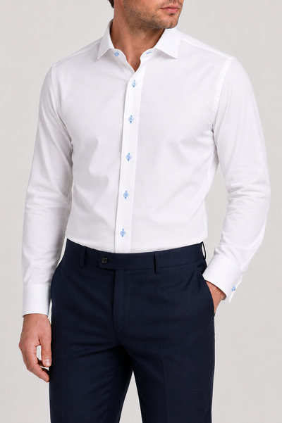 A man wearing the Wilkes & Riley White/Sky Spread Collar B1715 dress shirt, featuring a crisp white twill fabric with light blue buttons, pairs it with dark navy trousers. The refined spread collar from Wilkes & Riley adds sophistication to his polished look as he stands with one hand in his pocket against a plain background.