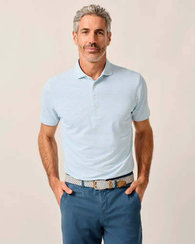 A man with gray hair and a beard stands with his hands in his pockets against a plain background, wearing blue pants and a Johnnie-O Performance Polo- Stetsons by Johnnie-O.