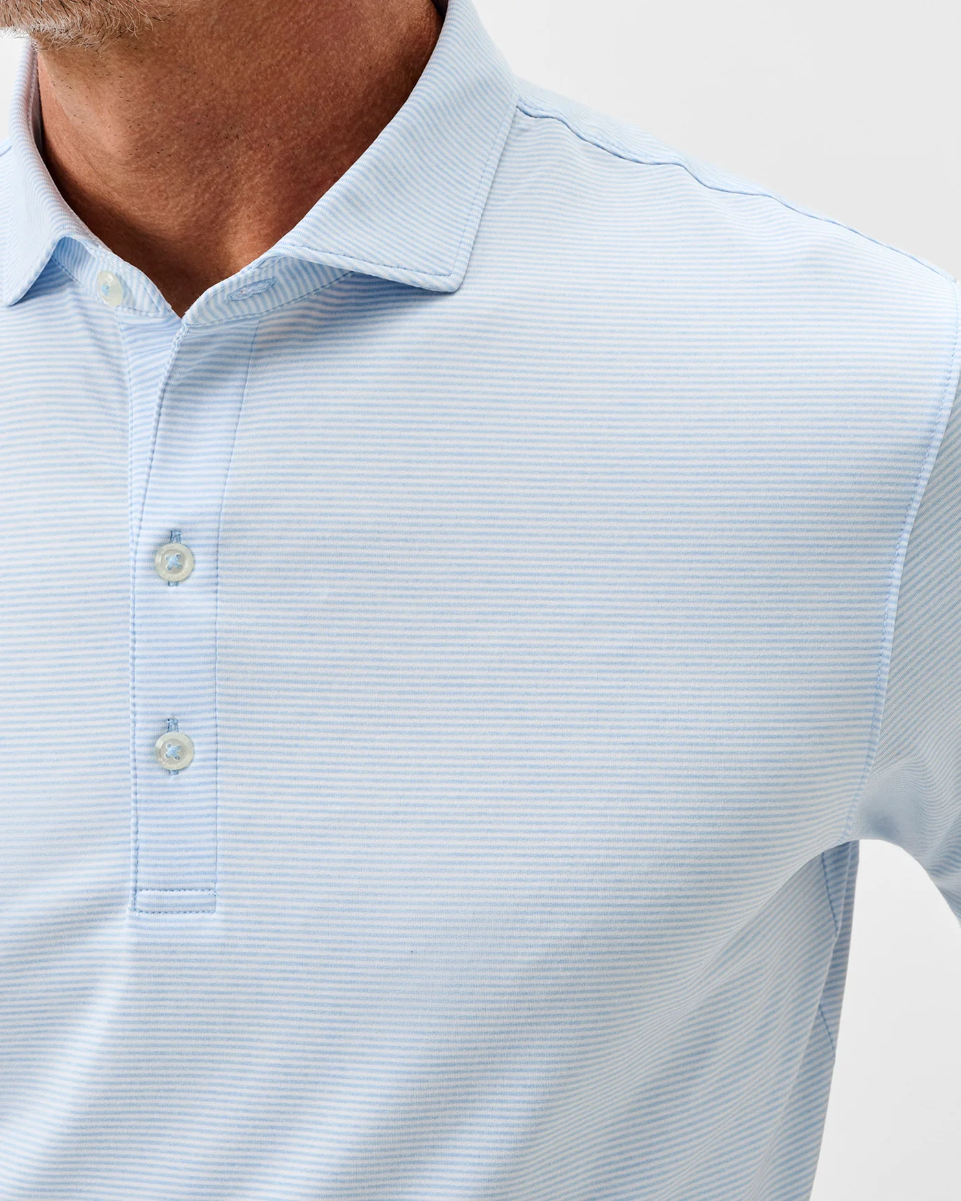 Close-up of a man wearing the Johnnie-O Lyndonn Stripe Performance Knit Polo, a casual light blue short sleeve shirt with thin horizontal stripes. Only the lower face, neck, and upper torso are visible. Made from breathable nylon-spandex blend.