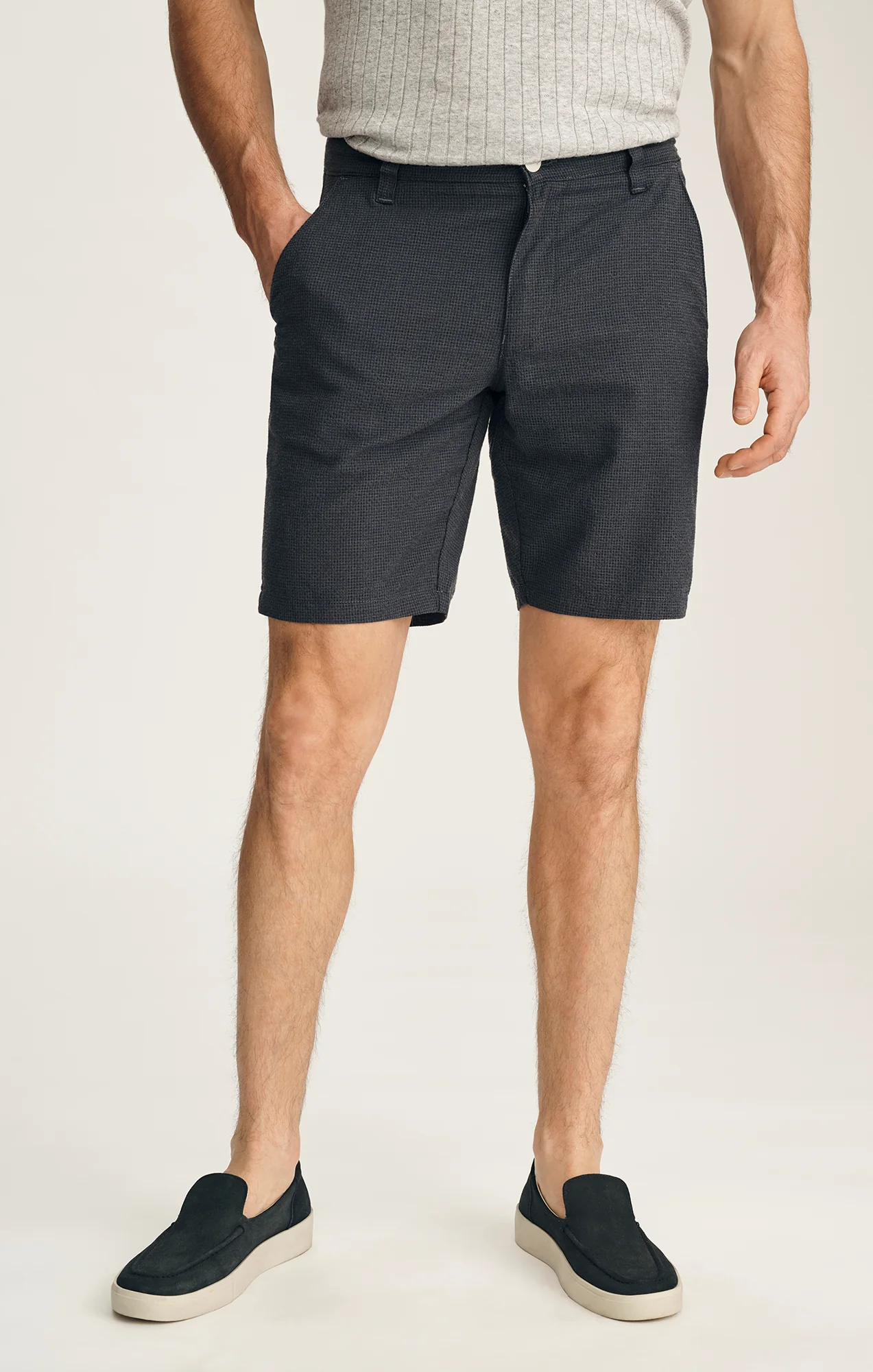 A man wearing a gray ribbed shirt, 34 Heritage Arizona Seersucker Shorts by 34 Heritage, and black slip-on shoes stands with hands in his pockets against a plain background.