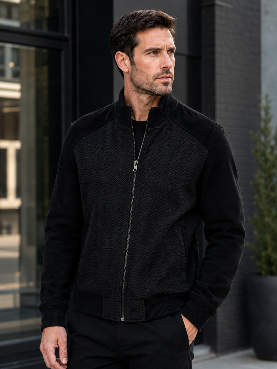 Wearing the Enzo Nino Suede Bomber Jacket by Enzo, a man stands outdoors in an urban setting with one hand in his pocket, gazing off to the side.